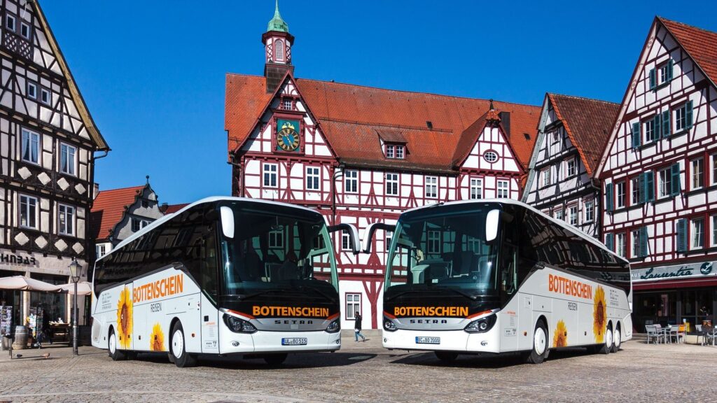 Zwei Reisebusse von Bottenschein Reisen vor historischen Fachwerkhaeusern.
